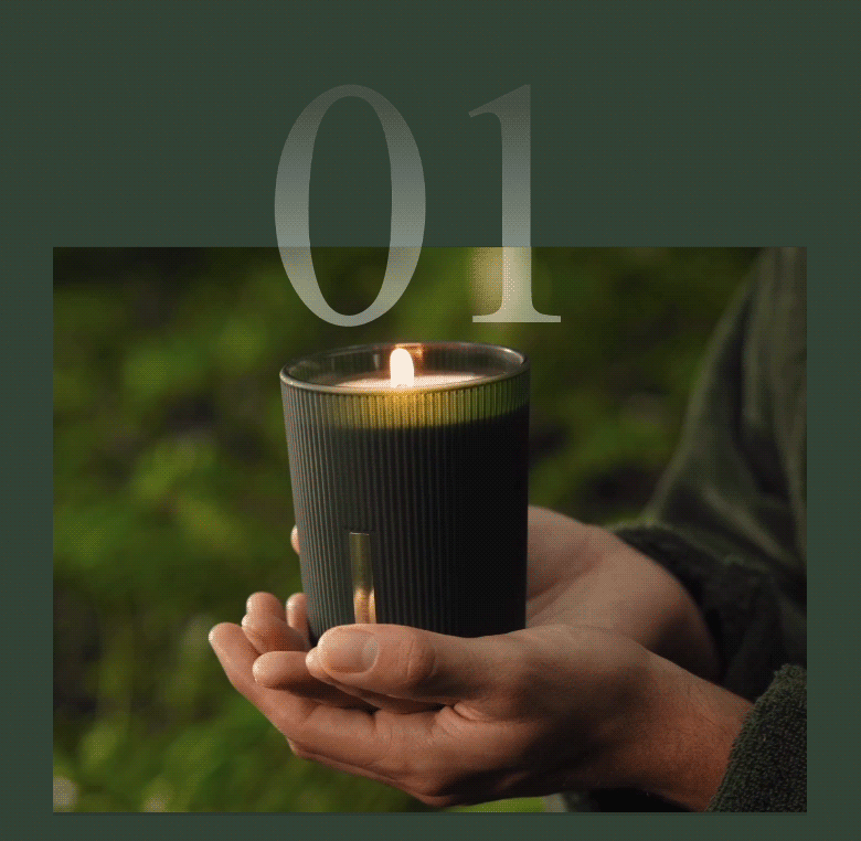 Hands cradling a lit scented candle in a dark green glass holder