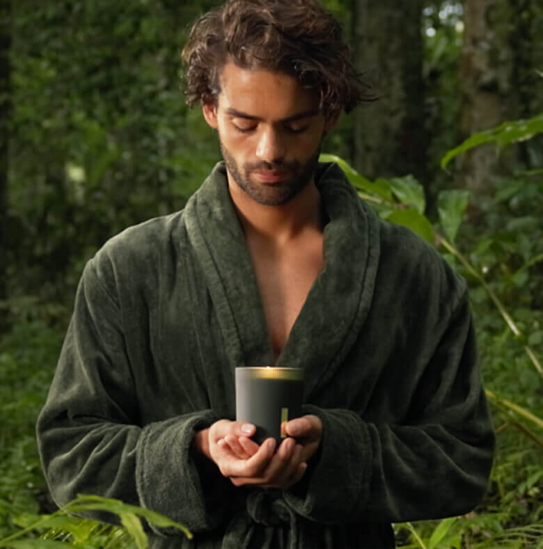 Person in green robe holding a scented candle in a lush forest setting